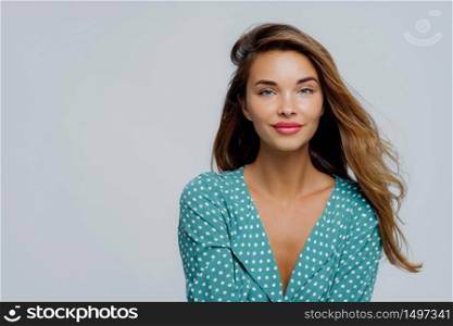 Studio shot of positive young woman has long wavy hair, makeup, wears turquoise polkadot blouse, looks straightly at camera, models against white background, copy space for your advertisement