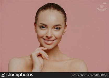 Studio shot of pretty charming woman touches chin, has healthy glowing skin, cares about complexion, stands naked indoor, has cosmetology procedure, isolated on pink background. Beauty concept