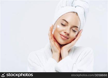 Studio shot of relaxed beautiful woman with closed eyes tilts head, touches skin gently, applies face cream, wears bath towel on head after taking spa procedures, enjoys facial treatments, body care