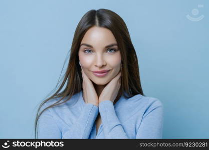 Studio shot of tender young woman touches cheeks, looks directly at camera with tender gaze, poses indoors, feels relaxed, listens something carefully, has natural beauty. Skin care concept.