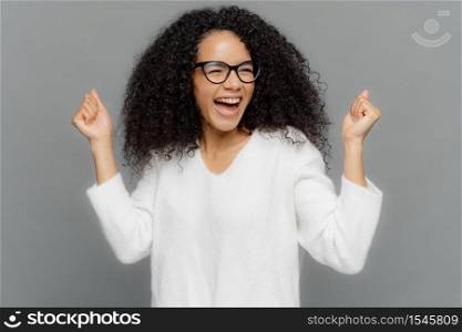 Studio shot of triumphing dark skinned woman clenches fists, smiles happily, celebrates success, achieves goal, smiles broadly, dressed casually, isolated over grey background. Victory concept