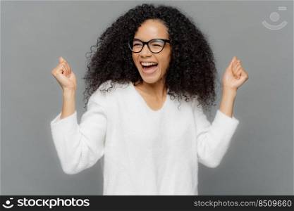 Studio shot of triumphing dark skinned woman clenches fists, smiles happily, celebrates success, achieves goal, smiles broadly, dressed casually, isolated over grey background. Victory concept