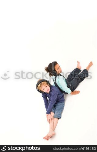 Studio shot of two young children, one a blond boy the other a mixed race girl, sitting back to back and listening to music on headphones. Studio shot on a white background isolated with a shadow for depth.