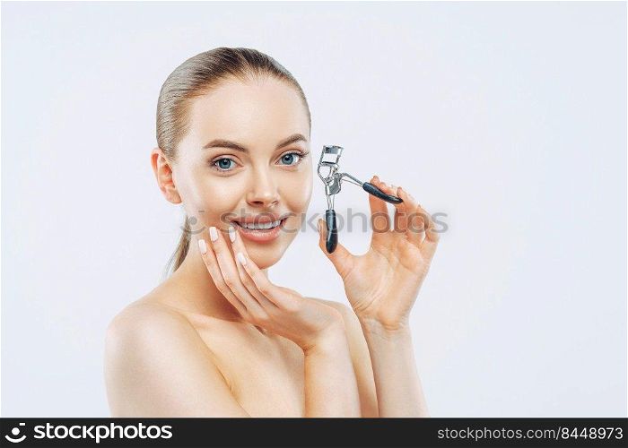 Studio shot of young brunette woman with tender smile, uses eyelash curler, has smooth soft skin, stands naked, has natural makeup, touches face, poses against white background. High resolution