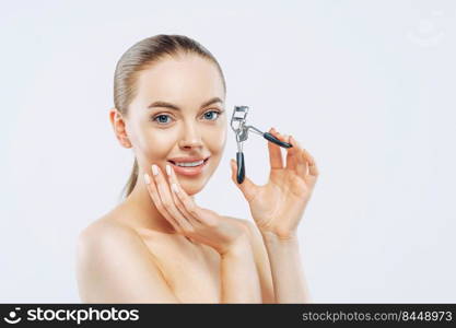 Studio shot of young brunette woman with tender smile, uses eyelash curler, has smooth soft skin, stands naked, has natural makeup, touches face, poses against white background. High resolution