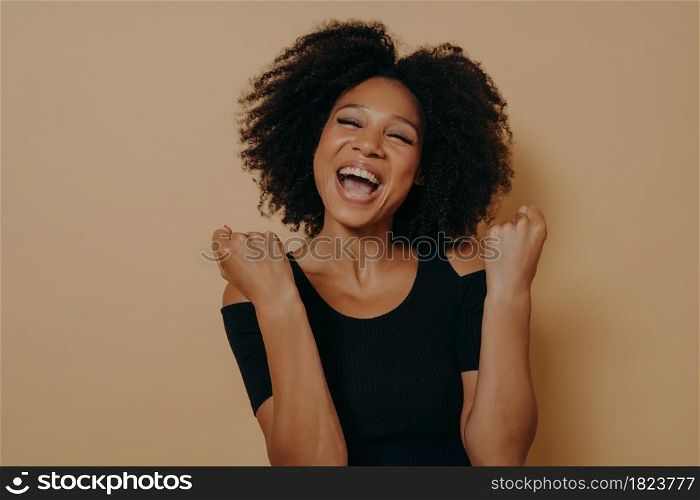Studio shot of young joyful excited dark skinned woman keeping fists clenched and eyes closed, screaming from enjoyment while posing against beige background. Success and triumph concept. Young joyful excited dark skinned woman keeping fists clenched while posing against beige background