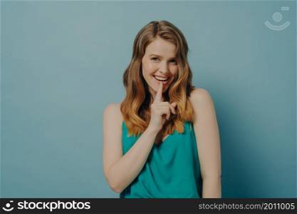 Studio waist-up portrait of flirty young woman shows silence sign with finger touching her lips, asking to keep your mouth shut, looking with seductive smile at camera, isolated over blue background. Flirty young woman shows silence Shhh symbol with index finger touching her lip, isolated on blue