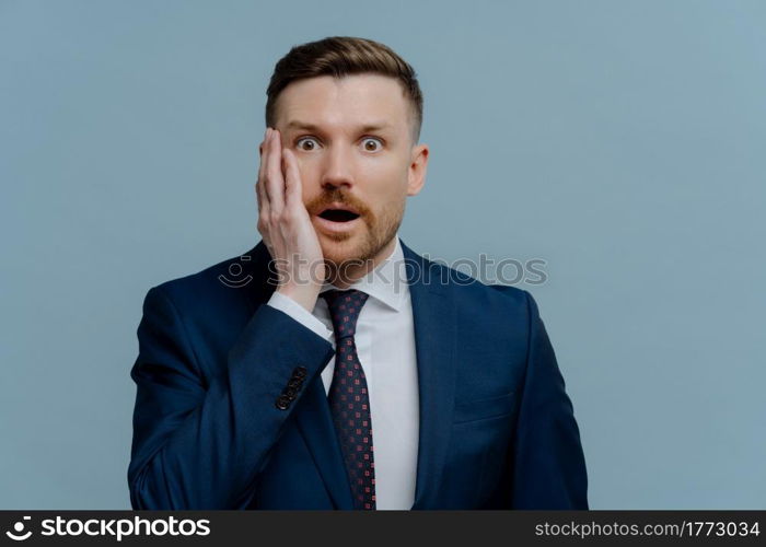 Stunned businessman in formal wear getting bad unpleasant news about his business, looking at camera with open mouth and shocked face expression while standing against light steel blue background. Surprised businessman receiving negative news and feeling shocked