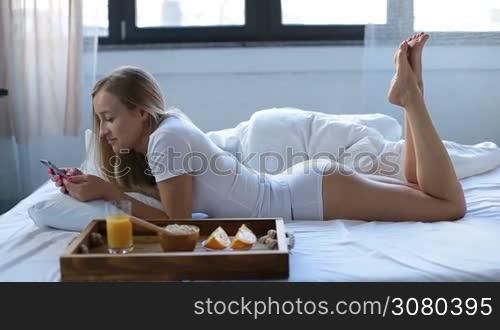 Stunning blonde woman in sexy white underwear lying on her belly in bed surfing the net with smartphone while having healthy breakfast early morning. Side view. Full length. Beautiful female relaxing on bed sheet and using mobile phone. Dolly shot.