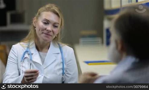 Stunning cheerful blonde female physician with stethoscope in lab coat holding eletronic thermometer and communicating with patient at clinic. Young serious female doctor checking body temperature of senoir ill man during medical exam.