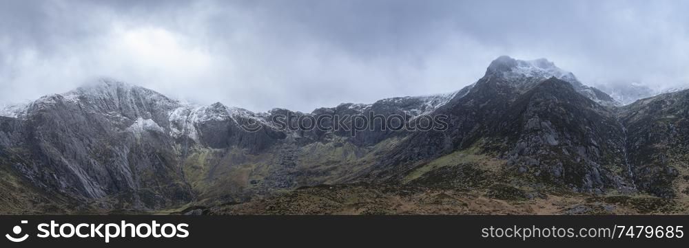 Stunning dramatic panorama landscape image of snowcapped Glyders mountain range in Snowdonia during Winter with menacing low cloudshanging at the mountain peaks