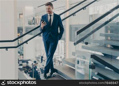 Stunning entrepreneur with neat beard in formal stylish suit standing on staircase in office lobby with smartphone in his hand, checking e-mails, financial reports or messages from business partners. Stunning entrepreneur in formal stylish suit standing on staircase in office lobby with smartphone
