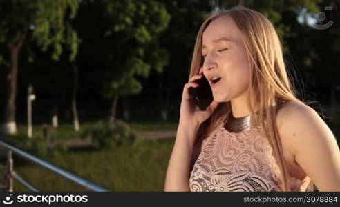 Stunning female in fashionable dress chatting on smartphone and smiling while leaning on railing on pedestrian bridge at sunset. Beautiful blond woman talking on smartphone in springtime against cityscape bakground. Slow motion. Stabilized shot.