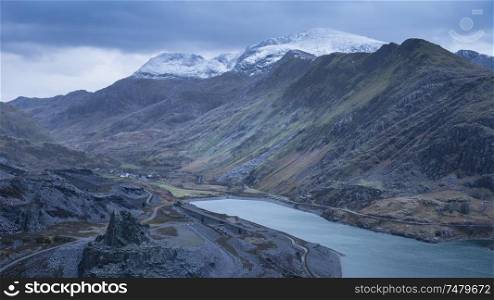 Stunning landscape image of Dinorwig Slate Mine and snowcapped Snowdon mountain in background during Winter in Snowdonia with Llyn Peris in foreground