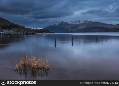 Stunning landscape image of Loch Lomond and snowcapped mountain range in distance viewed from small village of Luss