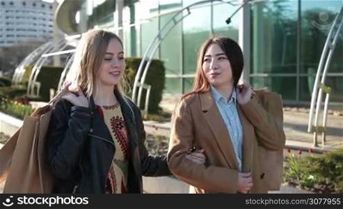 Stunning long hair brunette asian girl and charming blonde caucasian female holding shopping bags over their shoulders and walking along city street at sunset. Excited shopaholic women chatting about purchases after good shopping day.