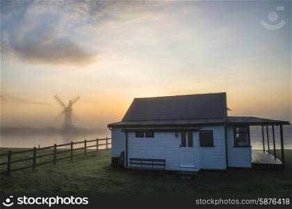 Stunning sunrise landscape over foggy river with windmill in background