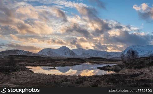 Stunning Winter panorama landscape image of mountain range viewed from Loch Ba in Scottish Highlands with dramatic clouds overhead
