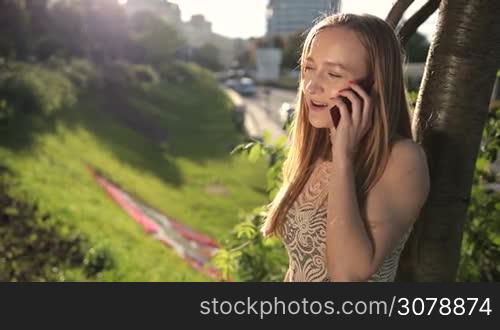 Stunning woman with amazing long blonde hair communicating on smartphone while leaning on the tree in the park. Charming lady talking on mobile phone outdoors in the flow of warm sunset. Slow motion. Steadicam stabilized shot.