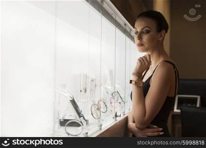 Stunning young brunette , watching jewel inside a jewellery , she stand near the shop window indoor .