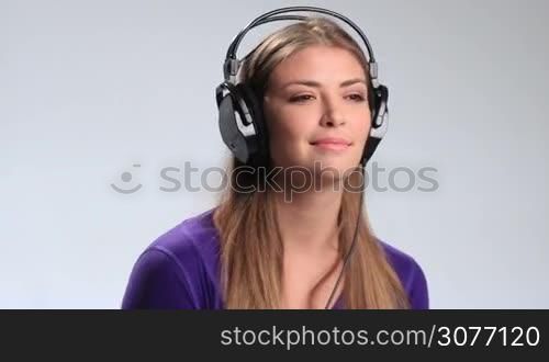 Stunning young brunette woman in earphones enjoying music on white. Cheerful teenage girl looking at camera silently saying what and laughing. Girl lifting one earcups with smile and noding then putting earcup back on ear and continues listening.