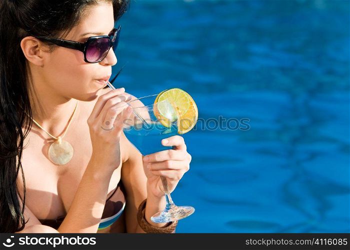 Stunningly beautiful young latina Hispanic woman drinking a cocktail by a blue swimming pool