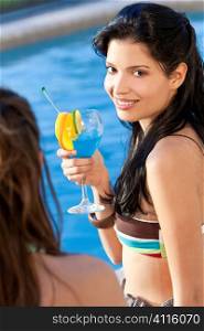 Stunningly beautiful young latina Hispanic woman in drinking a cocktail by a blue swimming pool with her friend in the foreground.