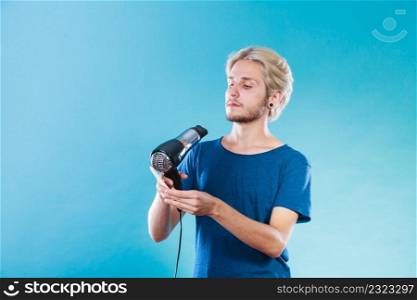 Style and fashion. Young trendy male hairstylist barber with new idea of look changing. Blonde man holding hair dryer and comb creating new hairdo, on blue. Trendy man with hair dryer