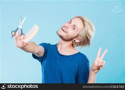 Style and fashion. Young trendy male hairstylist barber with new idea of look changing. Blonde guy with scissors and wooden comb creating hairdo coiffure, on blue. Man with scissors and comb creating new coiffure