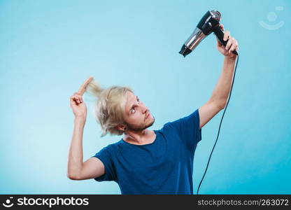 Style and fashion. Young trendy male hairstylist barber with new idea of look changing. Blonde man holding hair dryer and comb creating new hairdo, on blue. Trendy man with hair dryer