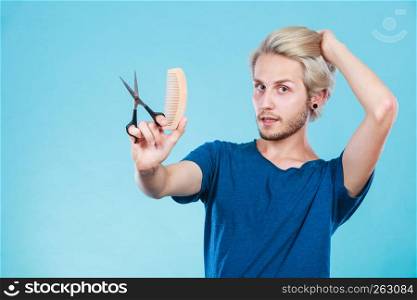 Style and fashion. Young trendy male hairstylist barber with new idea of look changing. Blonde guy with scissors and wooden comb creating hairdo coiffure, on blue. Man with scissors and comb creating new coiffure