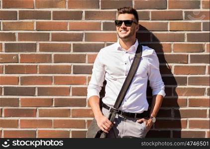 style and people concept - happy smiling young man in sunglasses with bag over brickwall. young man in sunglasses with bag over brickwall