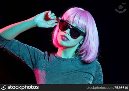 style, fashion and nightlife concept - happy young woman in pink wig and black sunglasses over neon light on dark background. happy woman in pink wig and black sunglasses