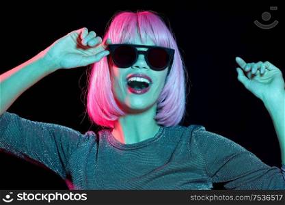 style, fashion and nightlife concept - happy young woman in pink wig and black sunglasses over neon light on dark background. happy woman in pink wig and black sunglasses