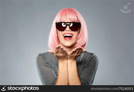 style, fashion and people concept - happy young woman in pink wig and black sunglasses holding something invisible on palms of her hands over grey background. happy woman in pink wig holding something on palms