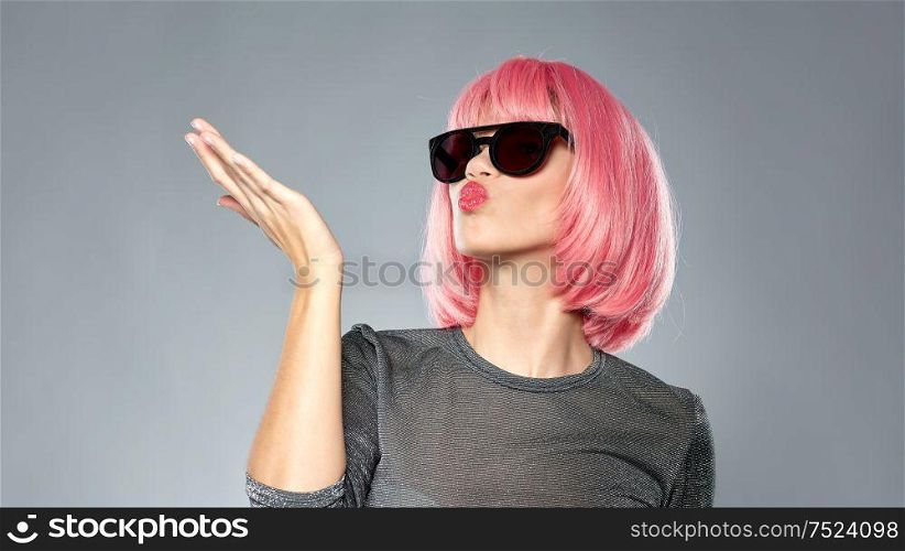 style, fashion and people concept - happy young woman in pink wig and black sunglasses sending air kiss to camera over grey background. woman in pink wig and sunglasses sending air kiss
