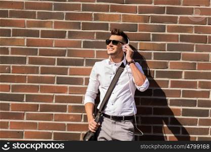 style, music, technology and people concept - happy smiling young man in headphones and sunglasses with bag over brickwall