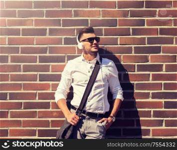 style, music, technology and people concept - happy smiling young man in headphones and sunglasses with bag over brickwall. young man in headphones with bag over brickwall
