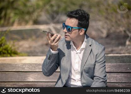 Stylish adult male entrepreneur in trendy suit and sunglasses communicating via voice message on mobile phone while sitting on wooden bench in summer day. Modern businessman recording voice message on smartphone