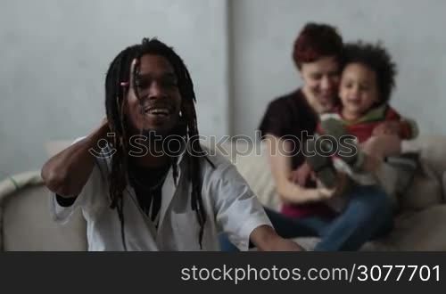 Stylish african american hipster father with dreadlocks sitting on the floor while his little mixed race toddler son coming to him from the back. Blurry cheerful caucasian mother sitting on the sofa. Slow motion