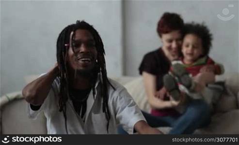 Stylish african american hipster father with dreadlocks sitting on the floor while his little mixed race toddler son coming to him from the back. Blurry cheerful caucasian mother sitting on the sofa. Slow motion