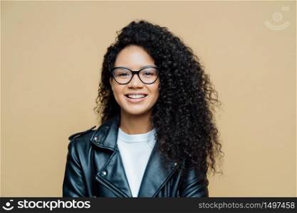 Stylish African American woman smiles toothily, has pleasant talk, enjoys good day, wears transparent glasses, leather jacket, expresses positive emotions, poses indoor at studio. Ethnicity, fashion