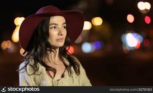 Stylish attractive long brown hair woman in fashionable hat looking at camera and smiling over defocused night city streetlights background. Beautiful positive female gazing at camera with radiant toothy smile in the street at night.