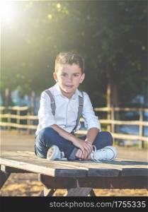 Stylish boy in suspenders and jeans sitting on table in park looking at camera.. Adorable trendy kid posing in park