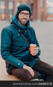 Stylish Caucasian man has free time, poses outside with disposable cup of coffee, dressed in hat and jacket, enjoys good rest, waits for someone at street, has satisfied expression. Lifestyle concept