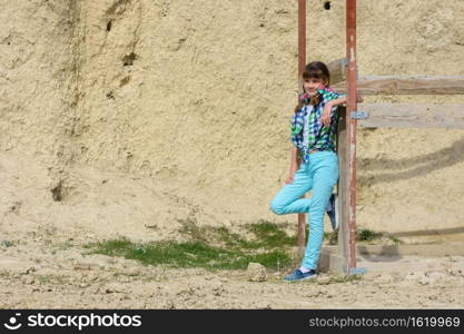 Stylish cute girl in a plaid shirt and jeans leans on a wooden structure against a background of a rock