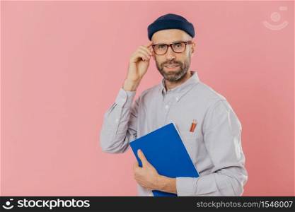 Stylish distant worker wears transparent glasses, stylish hat, shirt, holds blue textbook, uses pencils for writing information, poses over pink background. Satisfied male writer analyzes information
