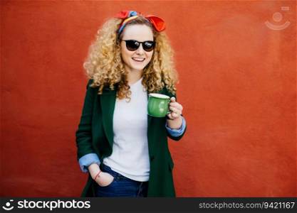Stylish European woman in headband, jacket, and shades, holding a cup of cappuccino. Happy expression, standing against orange background. People, lifestyle.
