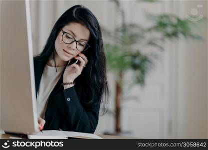 Stylish female entrepreneur in black suit, has phone conversation, makes notes in organizer looks assured and ambitious poses at desktop against office interior. Young woman manager talks via cellular