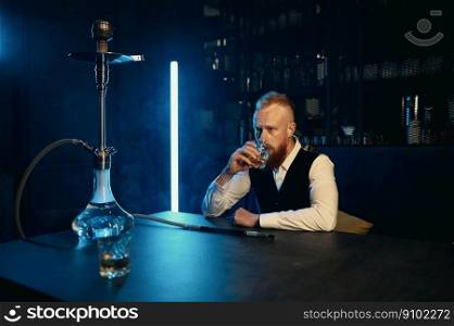 Stylish guy in suit drinking whisky from glass at hookah bar. Portrait of confident wealthy businessman. Stylish guy in suit drinking whisky at hookah bar